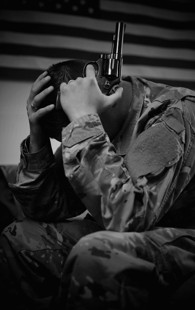 This is a black and white image of a soldier in full uniform holding a gun near his head.